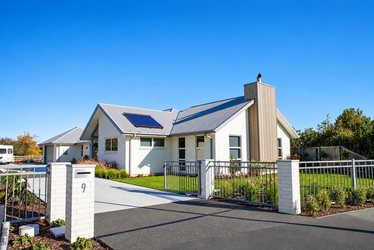 Une maison récente en briques blanches avec toit en tôle grise, panneau solaire et clôture métallique, sous un ciel bleu.