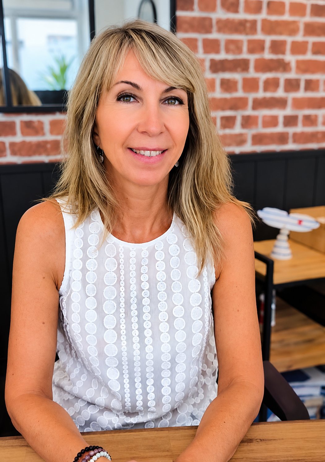 Une femme aux cheveux blonds mi-longs, vêtue d'un haut blanc sans manches, sourit assise à une table en bois devant un mur de briques rouges.