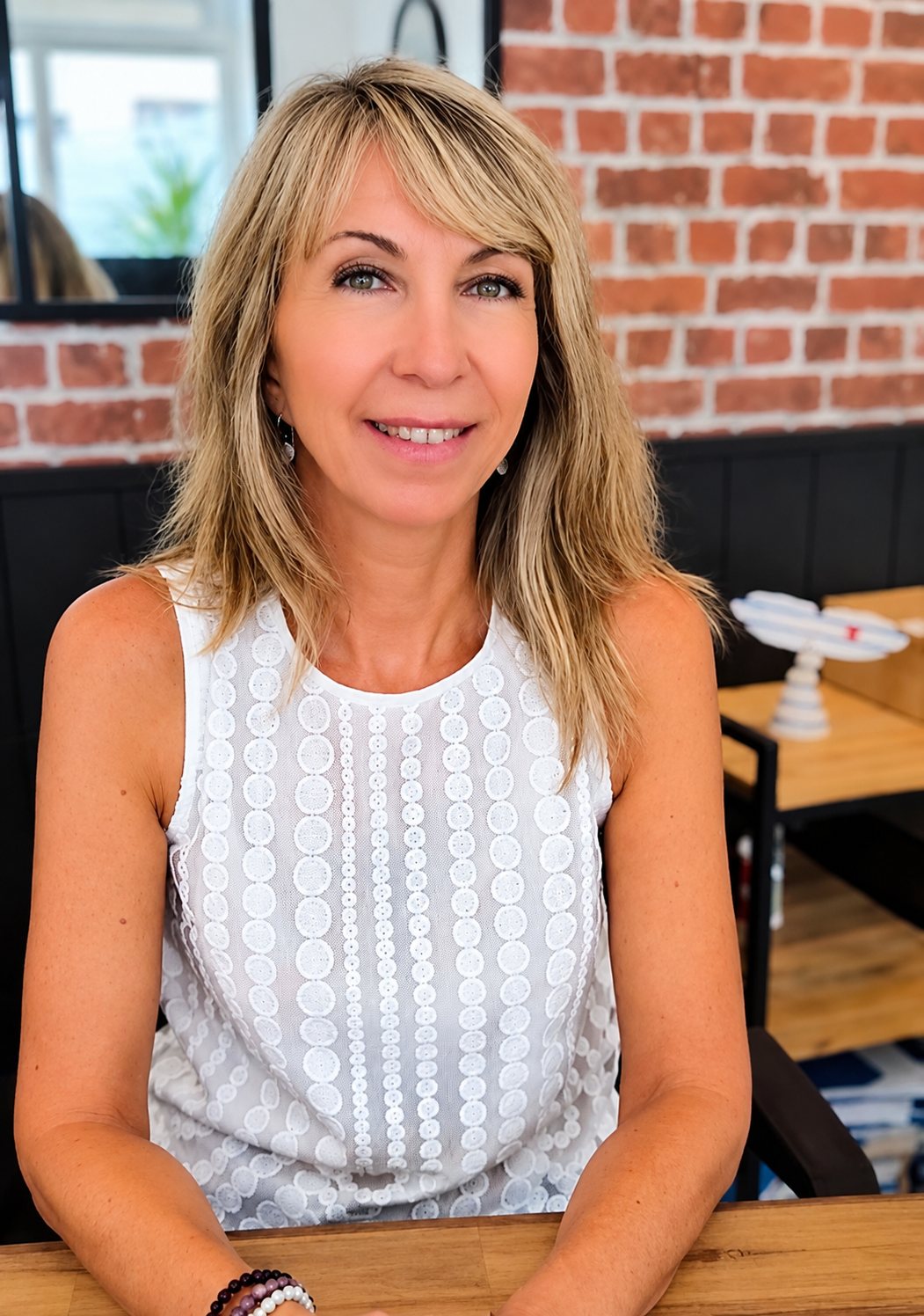 Une femme blonde aux cheveux mi-longs, vêtue d'un haut blanc sans manches, sourit, assise à une table en bois devant un mur de briques rouges.