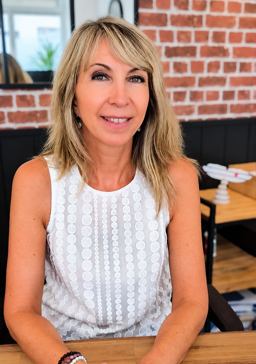 Une femme blonde aux cheveux mi-longs et en haut blanc brodé sourit, assise à une table en bois devant un mur de briques rouges.