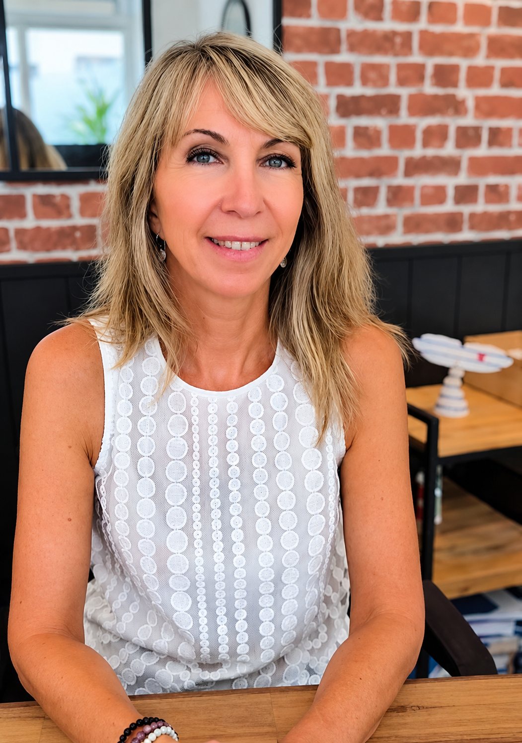 Une femme blonde aux cheveux mi-longs, vêtue d'un haut blanc sans manches, sourit assise à une table en bois devant un mur de briques.