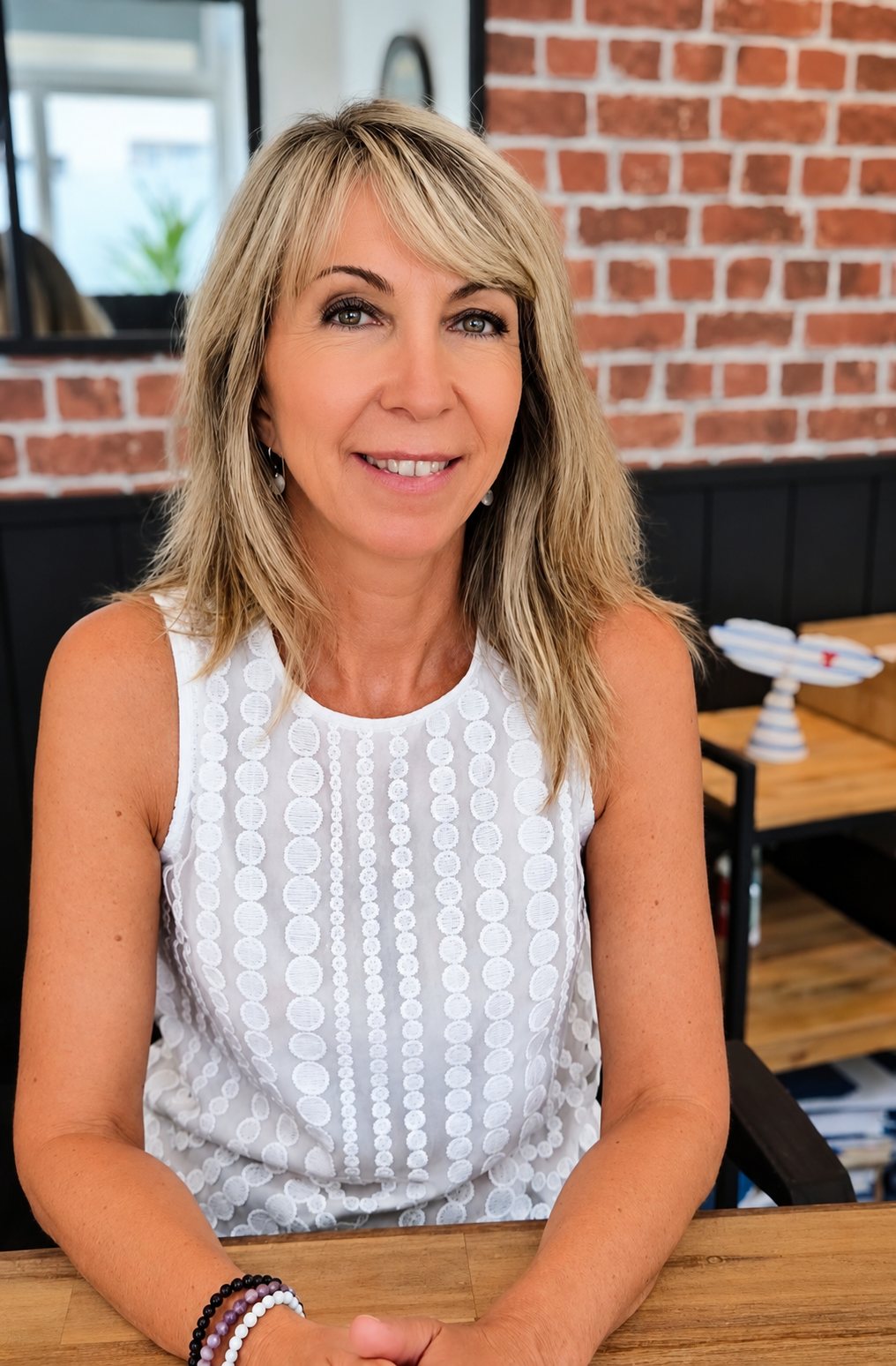 Une femme blonde aux cheveux mi-longs, vêtue d'un haut blanc sans manches, sourit à une table en bois devant un mur de briques rouges.